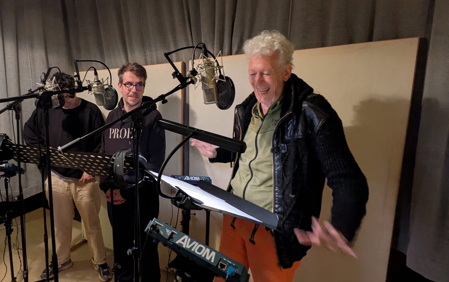 Dita steht am Studiomikrofon und performt als Holle. Tommy schaut ihr lächelnd zu.