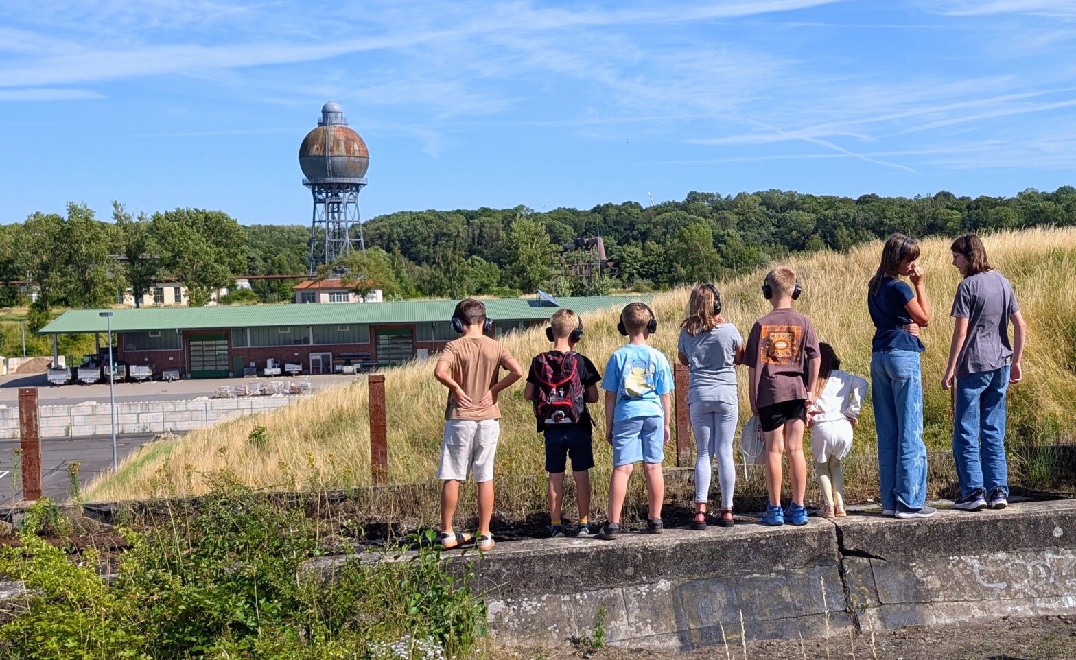 Sieben Kinder stehen auf einer Mauer und blicken auf das Gelände mit dem Kugelwasserturm im Hintergrund.